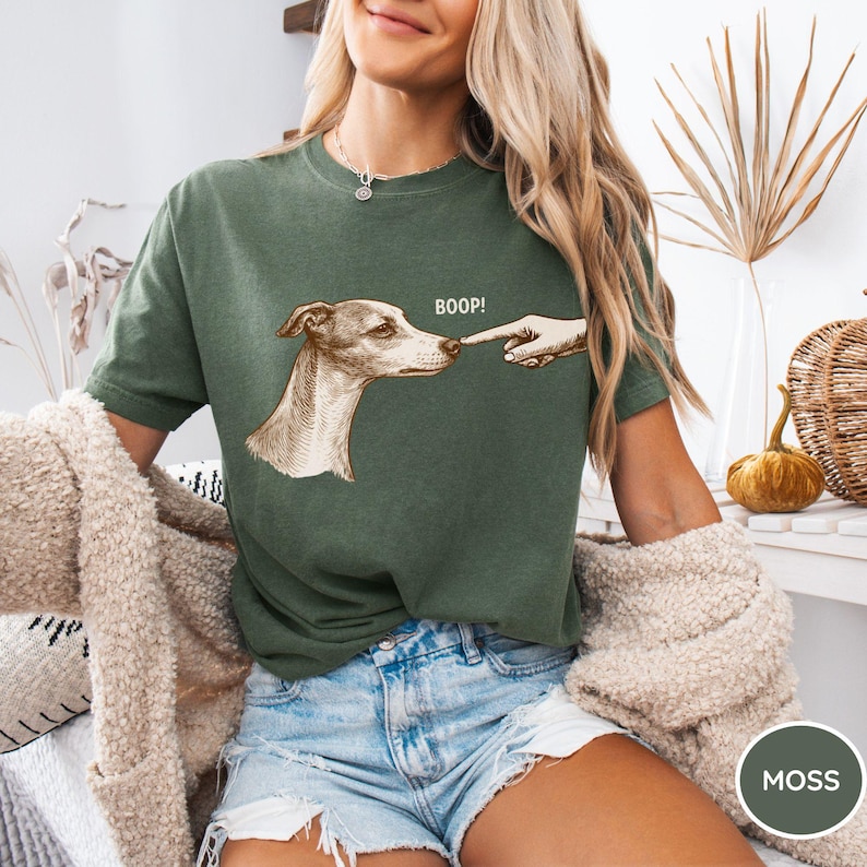 May include: A moss green short-sleeved t-shirt featuring a graphic illustration of a dog being touched on the nose by a human finger. The text BOOP! is printed in white above the dog's head. The shirt has a crew neckline and a relaxed fit.