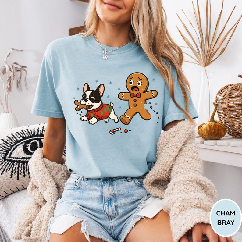 May include: Light blue t-shirt featuring a cartoon design of a French bulldog in a Christmas sweater chasing a gingerbread man. The gingerbread man has a scared expression. A candy cane is on the ground. The shirt has the text "CHAM BRAY" on the bottom right.