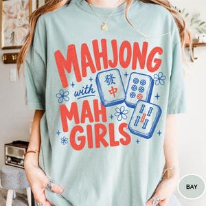 May include: A light blue t-shirt featuring red text that reads MAHJONG with MAH GIRLS. The design includes blue illustrations of three mahjong tiles and small flower accents. The shirt has a crew neckline and short sleeves, showcasing a graphic print.