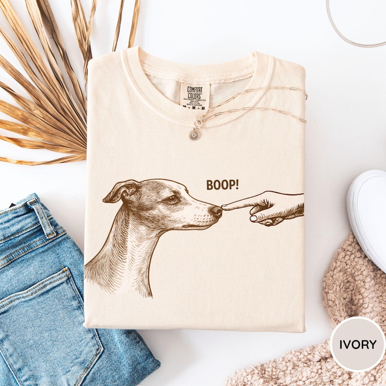 May include: An ivory-colored crew neck t-shirt featuring a brown line-art illustration of a dog being touched on the nose by a finger, with the text BOOP! printed above the dog's snout. The shirt has a Comfort Colors brand label at the neck.