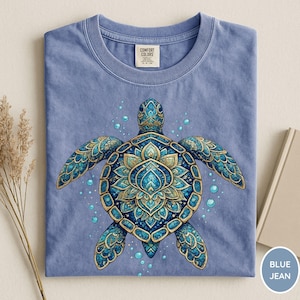 May include: A blue jean colored t-shirt featuring a central graphic of a sea turtle with a detailed, ornate shell pattern in shades of blue and gold. The turtle is surrounded by small bubbles. The shirt has a Comfort Colors brand neck label.