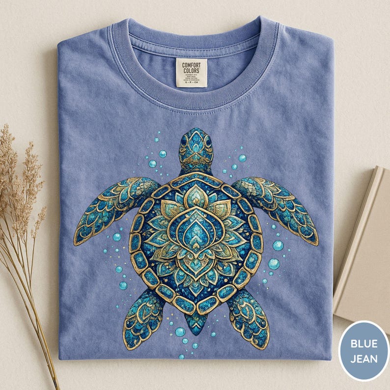 May include: A blue jean colored t-shirt featuring a central graphic of a sea turtle with a detailed, ornate shell pattern in shades of blue and gold. The turtle is surrounded by small bubbles. The shirt has a Comfort Colors brand neck label.