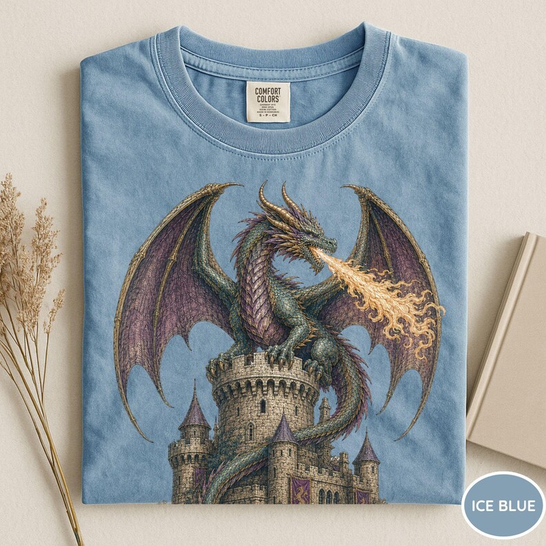 May include: A light blue t-shirt featuring a graphic print of a green dragon with purple wings perched atop a stone castle tower, breathing a stream of yellow fire. The shirt has a crew neckline and a Comfort Colors brand label at the collar.