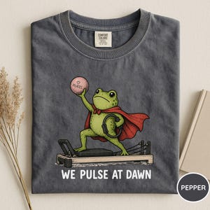 May include: A charcoal gray t-shirt featuring a graphic of a green frog wearing a red cape, standing on a pilates reformer machine. The frog holds a pink ball labeled "PILATES" and a black ring. The text "WE PULSE AT DAWN" is printed below.