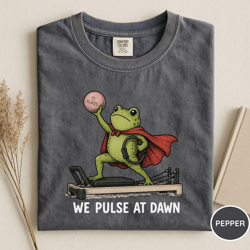 May include: A charcoal gray t-shirt featuring a graphic of a green frog wearing a red cape, standing on a pilates reformer machine. The frog holds a pink ball labeled "PILATES" and a black ring. The text "WE PULSE AT DAWN" is printed below.