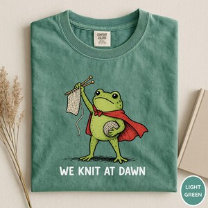 May include: Light green t-shirt featuring a cartoon frog superhero with a red cape, holding knitting needles and yarn. The text "WE KNIT AT DAWN" is printed below the frog. The shirt has a crew neck and short sleeves.