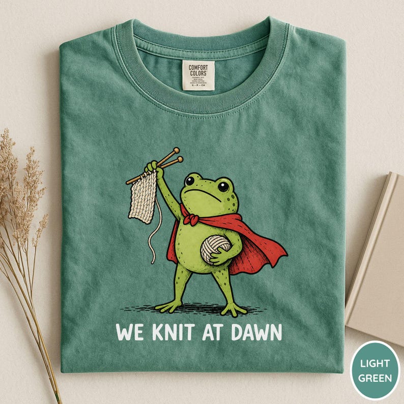 May include: Light green t-shirt featuring a cartoon frog superhero with a red cape, holding knitting needles and yarn. The text "WE KNIT AT DAWN" is printed below the frog. The shirt has a crew neck and short sleeves.