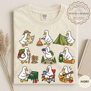 May include: An ivory t-shirt featuring a graphic print of nine white ducks engaged in various camping activities, including reading a map, roasting a marshmallow, setting up a tent, hiking, using binoculars, holding a lantern, cooking, and playing guitar.