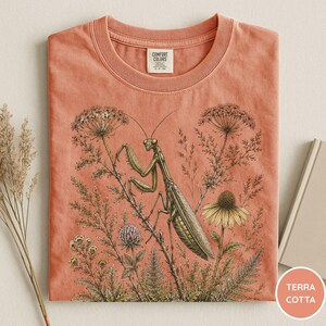 May include: Terracotta-colored t-shirt featuring a detailed illustration of a praying mantis amidst wildflowers and foliage. The shirt has a crew neck and a tag that reads "Comfort Colors". The design includes a variety of plants and flowers.