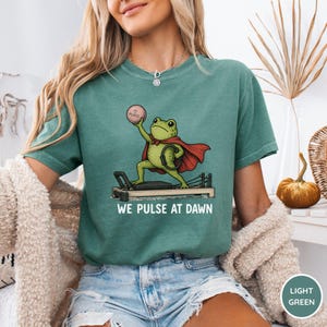 May include: A light green t-shirt featuring a graphic of a green frog wearing a red cape while exercising on a pilates reformer machine. The frog holds a pink ball labeled Pilates. Below the graphic, the text reads WE PULSE AT DAWN.