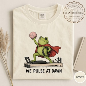 May include: An ivory t-shirt featuring a graphic of a green frog wearing a red cape, standing on a black pilates reformer machine. The frog holds a pink ball labeled Pilates and a black ring. The text below reads WE PULSE AT DAWN.