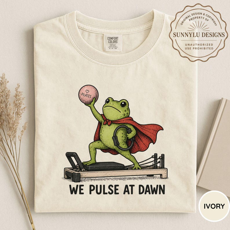 May include: An ivory t-shirt featuring a graphic of a green frog wearing a red cape, standing on a black pilates reformer machine. The frog holds a pink ball labeled Pilates and a black ring. The text below reads WE PULSE AT DAWN.