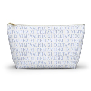 May include: A white cosmetic bag with a repeating pattern of the words "ALPHA XI DELTA" in light blue. The bag has a gold zipper and is designed to hold makeup or toiletries.