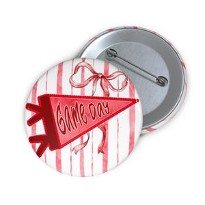 May include: A round button with a red and white striped background. The button features a red pennant with the words "Game Day" and a red bow. The button is a fun accessory for sports fans.