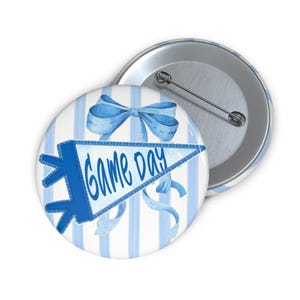 May include: A round button with a blue and white striped background. The button features a blue pennant with the words "GAME DAY" and a blue bow. The button is made of metal and has a pin on the back.