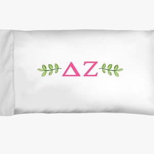 May include: White pillowcase with pink Greek letters Delta and Zeta, flanked by green leaf sprigs. The pillowcase is rectangular and made of a smooth, white fabric. The design is centered on the pillowcase.