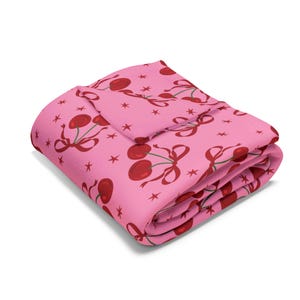 Puede incluir: Manta de forro polar rosa con un patrón repetido de cerezas rojas con tallos verdes y lazos rosas. La manta está doblada en tres partes y tiene un fondo blanco.