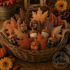 May include: A woven basket filled with fabric maple leaves and stuffed fabric acorns in various patterns and colors including brown, orange, and green. The leaves feature herringbone, striped, and plaid prints. A circular logo reads Mountain Range Farms.