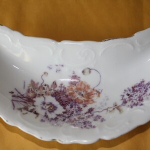 May include: A white porcelain half-moon shaped dish with a floral design in shades of purple and pink. The dish has a gold rim and is decorated with a delicate floral pattern.