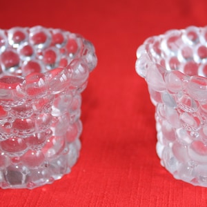 May include: Two clear glass candle holders with a textured, bubble-like design. The holders are cylindrical and have a wide opening at the top.