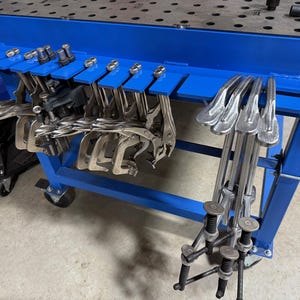 Puede incluir: Un carro de herramientas de metal azul con varias abrazaderas y herramientas. El carro tiene un marco azul y una parte superior con múltiples agujeros. Varias abrazaderas cuelgan de la parte superior y otras herramientas están de pie. El carro está sobre ruedas.