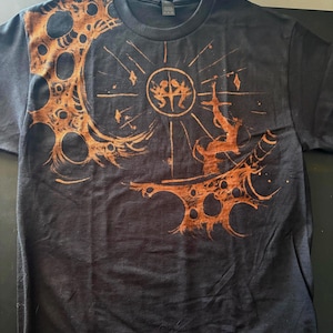 Puede incluir: Camiseta gris oscuro con un dise&ntilde;o gr&aacute;fico naranja. El dise&ntilde;o incluye un elemento circular con figuras, una forma de media luna con recortes circulares y una forma de espada. La camiseta tiene cuello redondo.