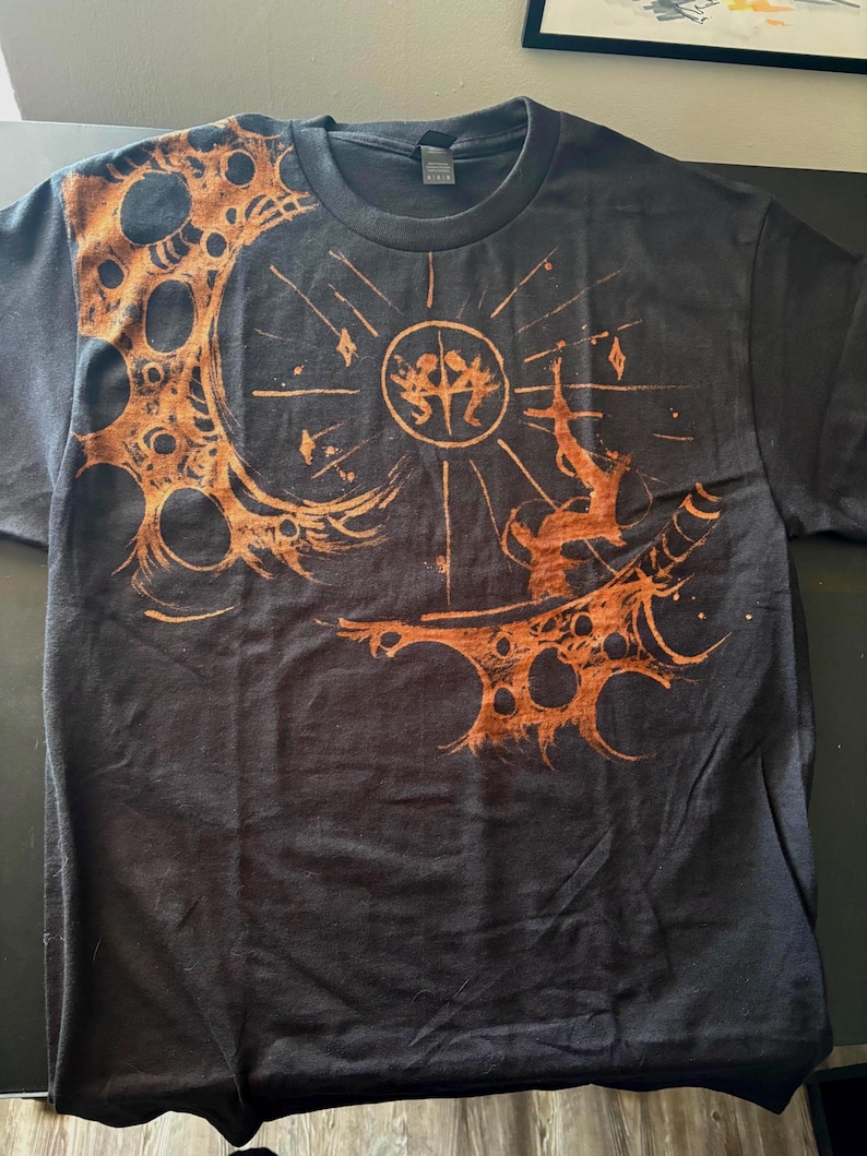 Puede incluir: Camiseta gris oscuro con un dise&ntilde;o gr&aacute;fico naranja. El dise&ntilde;o incluye un elemento circular con figuras, una forma de media luna con recortes circulares y una forma de espada. La camiseta tiene cuello redondo.