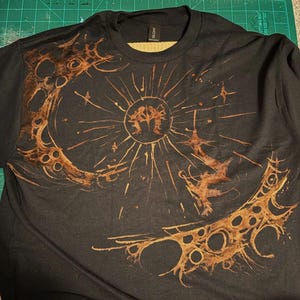 Puede incluir: Camiseta negra con un dise&ntilde;o de sol y luna en tonos marrones. El sol tiene l&iacute;neas radiantes y una figura central, mientras que la luna es creciente con recortes circulares. El dise&ntilde;o tiene un aspecto desgastado y art&iacute;stico.
