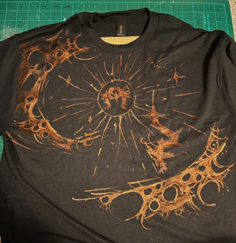 Puede incluir: Camiseta negra con un dise&ntilde;o de sol y luna en tonos marrones. El sol tiene l&iacute;neas radiantes y una figura central, mientras que la luna es creciente con recortes circulares. El dise&ntilde;o tiene un aspecto desgastado y art&iacute;stico.