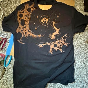 Puede incluir: Camiseta negra con un dise&ntilde;o intrincado en marr&oacute;n. El dise&ntilde;o presenta un emblema central, elementos celestiales y patrones detallados. La camiseta tiene cuello redondo y mangas cortas, ideal para uso casual.