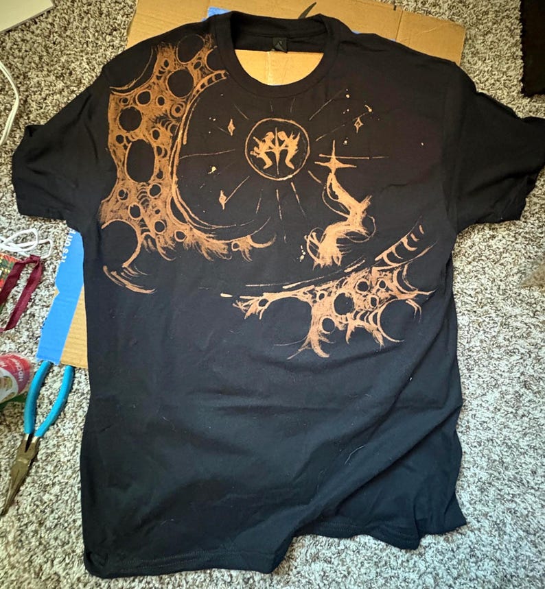 Puede incluir: Camiseta negra con un dise&ntilde;o intrincado en marr&oacute;n. El dise&ntilde;o presenta un emblema central, elementos celestiales y patrones detallados. La camiseta tiene cuello redondo y mangas cortas, ideal para uso casual.