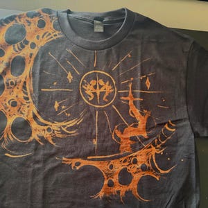 Puede incluir: Camiseta gris oscuro con un dise&ntilde;o gr&aacute;fico naranja y marr&oacute;n. El dise&ntilde;o presenta un sol con figuras, una forma de media luna y una figura que se extiende hacia arriba. La camiseta es de material suave.