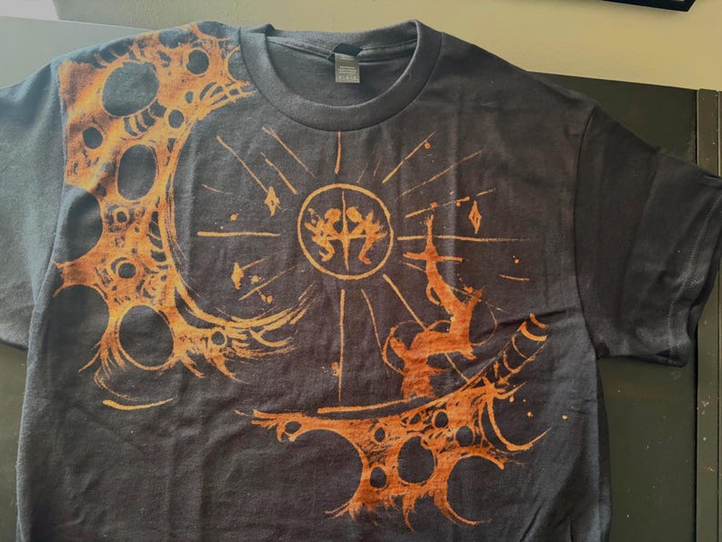 Puede incluir: Camiseta gris oscuro con un dise&ntilde;o gr&aacute;fico naranja y marr&oacute;n. El dise&ntilde;o presenta un sol con figuras, una forma de media luna y una figura que se extiende hacia arriba. La camiseta es de material suave.