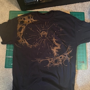 Puede incluir: Camiseta negra con un dise&ntilde;o circular marr&oacute;n. El dise&ntilde;o presenta una figura central con l&iacute;neas radiantes y un patr&oacute;n texturizado similar a la luna. La camiseta est&aacute; colocada sobre una esterilla de corte verde.