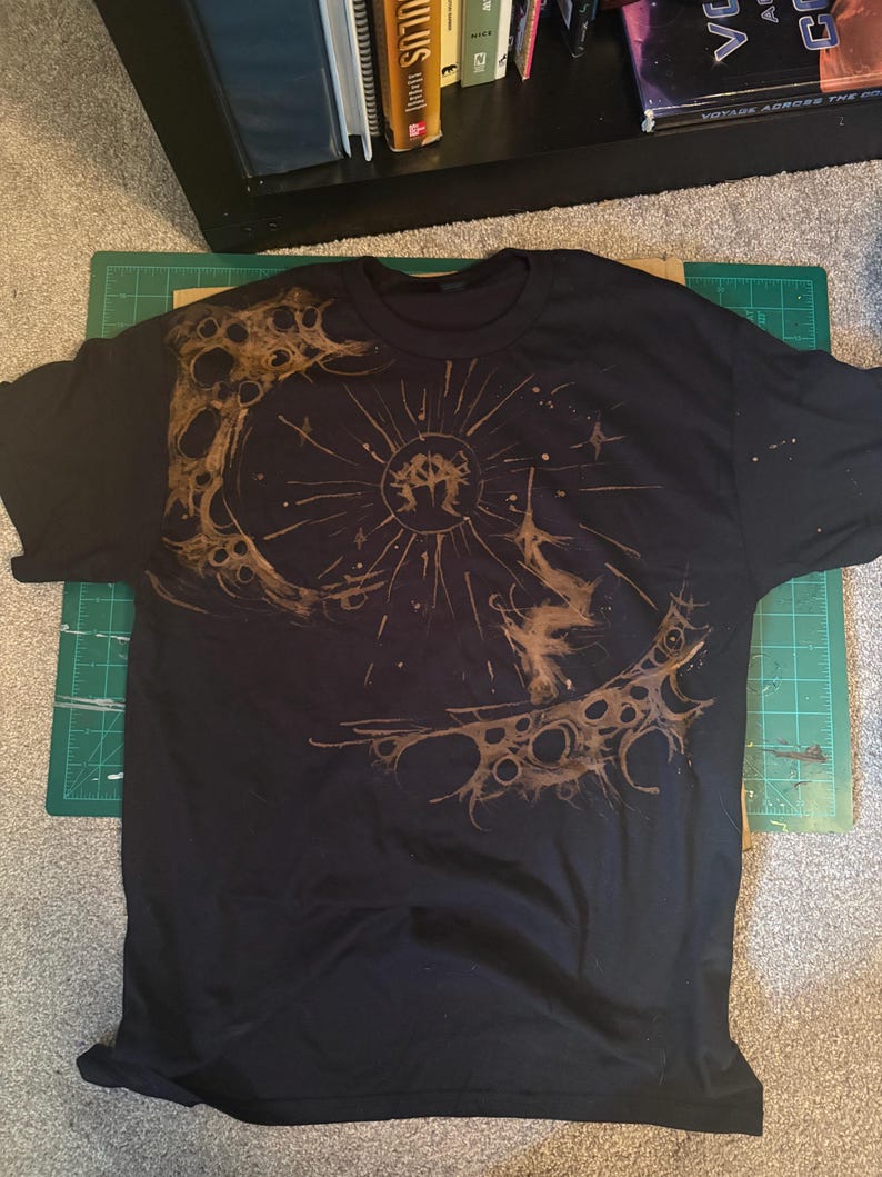 Puede incluir: Camiseta negra con un dise&ntilde;o circular marr&oacute;n. El dise&ntilde;o presenta una figura central con l&iacute;neas radiantes y un patr&oacute;n texturizado similar a la luna. La camiseta est&aacute; colocada sobre una esterilla de corte verde.