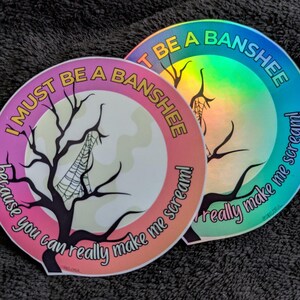 May include: Two circular stickers featuring a black silhouette of a bare tree with a spiderweb. The text reads I MUST BE A BANSHEE because you can really make me scream! One sticker has a pink and orange gradient border, while the other has a holographic finish.