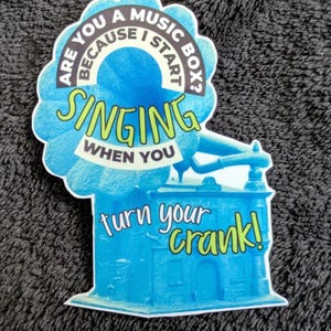 May include: A die-cut sticker featuring a bright blue vintage phonograph with a large horn. The text reads: ARE YOU A MUSIC BOX? BECAUSE I START SINGING WHEN YOU turn your crank! The design uses a mix of white, black, and lime green lettering.