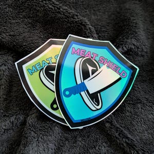 May include: Two iridescent stickers shaped like shields with the text "MEAT SHIELD" in black and white. The shield on the left is green and the shield on the right is blue.