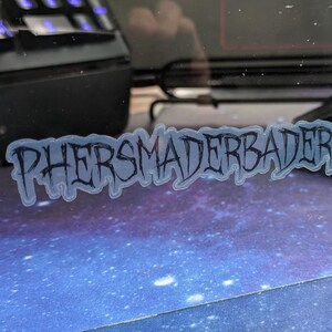 May include: A sticker with the word "PHERSMADERBADER" in a gothic font. The sticker has a light blue outline and a dark blue font. The sticker is on a blue surface with white specks.