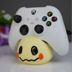 Puede incluir: Un mando blanco de Xbox descansa sobre un personaje amarillo en forma de huevo con una cara sonriente negra y detalles en naranja. El mando tiene joysticks negros y botones de colores.