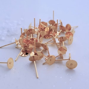 Könnte beinhalten: Ein Haufen goldfarbener Ohrring-Stifte mit flachen, runden Rückseiten. Die Stifte sind etwa 1 cm lang.