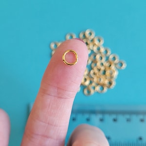 May include: A close-up of a small, gold-coloured jump ring on a fingertip, with a pile of similar rings blurred behind. The ring is approximately 0.6 cm in diameter. The background is a solid teal colour.