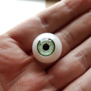 Puede incluir: Un ojo de cristal realista con iris verde claro y pupila negra. El ojo est&aacute; montado en una base blanca y redondeada. El ojo se sostiene en una mano, mostrando su detalle y artesan&iacute;a.