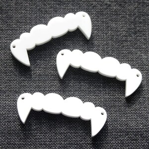 May include: Three white, flat, vampire fang-shaped charms with scalloped top edges and pointed ends. Each charm features two small circular holes, one at each pointed end, designed for attachment. The pieces have a smooth, solid white finish.