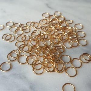 Puede incluir: Una colección de pequeños anillos de salto dorados esparcidos sobre una superficie de mármol blanco y gris. Los anillos son circulares y parecen ser de metal, probablemente para la fabricación de joyas o proyectos de artesanía.