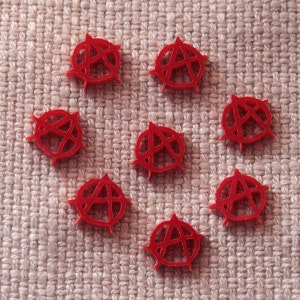 May include: Eight red anarchy symbol charms, each with a circle around the symbol. The charms are arranged in a heart shape on a beige fabric background.