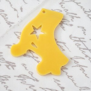 May include: A bright yellow, flat, die-cut acrylic charm in the shape of a stylized letter A. The letter features a small circular hole at the top for hanging and a five-pointed star cutout positioned within the upper section of the letter form.