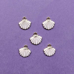 May include: Five white seashell charms with gold-colored trim and a loop for hanging. The charms are arranged on a purple background. These charms are ideal for jewelry making or other craft projects.