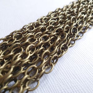 May include: Close-up of multiple strands of antique brass-coloured chain. The oval links are tightly interlocked, creating a textured surface. The chains are arranged diagonally across a white background, highlighting the metallic sheen and intricate design.