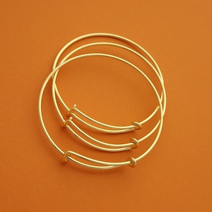 Puede incluir: Tres pulseras de oro apiladas una encima de la otra. Las pulseras son simples y tienen un acabado liso.
